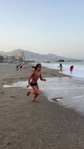 A young person with long brown hair runs along a sandy beach toward the ocean, wearing dark swim trunks. Their body is angled to the right, with their left leg extended forward and their arms slightly bent. The person's feet are bare, and footprints are visible in the sand. The ocean is to the left, with waves breaking and foam washing onto the shore. In the background, two other people are visible further down the beach, one in pink and the other in red. In the distance, buildings and mountains are visible along the coastline. The sky is a pale blue, transitioning to a lighter shade near the horizon. The video appears to be shot during the day, possibly late afternoon or early evening.