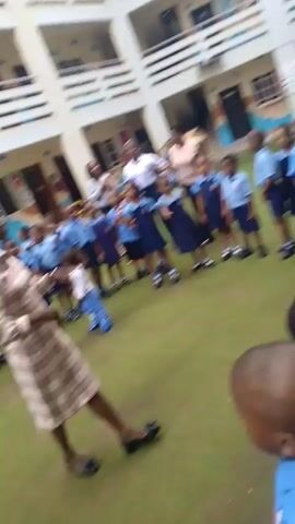 The video depicts an outdoor scene at a school or childcare facility, likely in Idimu, Nigeria, during daytime. A group of young children, predominantly dressed in blue uniforms (some with light blue shirts and dark blue shorts/skirts, others in full blue dresses), are arranged in a large circle on a green artificial turf area. Several adult women, presumably teachers, are present, supervising the children.

A prominent adult woman in a plaid dress and dark shoes stands within the circle, actively interacting with the children and appearing to lead an activity. She is seen moving around the circle and gesturing. The children are engaged, with some smiling, and appear to be participating in a structured game or movement activity, possibly to music heard in the background.

The setting includes a multi-story building in the background with balconies and decorative elements on its walls, consistent with school architecture. The weather appears clear and bright. No discernible text is visible within the frame.