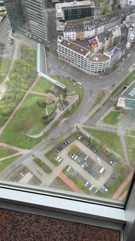 The video shows an aerial view of a city with multiple buildings, roads, and green spaces. The camera pans slowly to the left, revealing a broader cityscape. A tall, glass-walled building with a distinctive arch-shaped opening is visible in the lower left corner of the frame. Roads with moving cars and parked vehicles are seen, and various architectural styles of buildings are visible. The overall tone is observational and objective.
