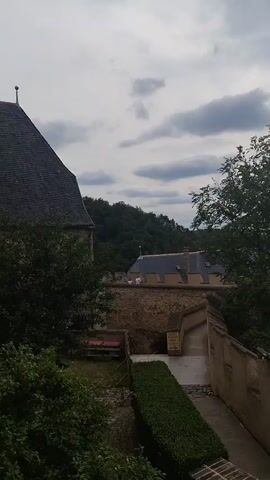 The video captures a scenic view from a stone wall overlooking a lush green landscape and a cloudy sky. In the foreground, a wall with crenellations forms a barrier. People are visible walking down a stone pathway, descending from the viewpoint. A man in a blue shirt and a dark baseball cap is seen holding up a smartphone, seemingly taking a picture or recording a video of the surrounding scenery. Further down the path, two children and an adult are interacting, with one child pointing towards the landscape. The environment appears to be a historical site, possibly a castle or fortress, given the architectural style of the walls and the distant building with a pointed roof. The foliage is dense, indicating a natural, possibly forested, area. The overall atmosphere is one of tourism and appreciation of natural beauty and historical architecture.