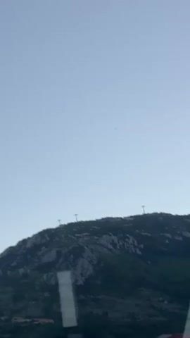 The video shows a view of a rocky, tree-covered hilltop against a clear, light blue sky. Three utility poles are visible along the ridge of the hill, connected by wires. A portion of a building, likely viewed from inside, is in the foreground, slightly obstructing the bottom left corner of the frame. The camera appears to be in motion, possibly from a vehicle.
