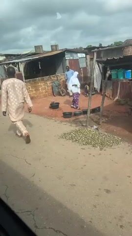 A video taken from a vehicle shows a street scene with several structures and people. The first structure is a building with a corrugated metal roof, brick walls, and a wooden frame. Next, a market stall is visible with a person standing behind it and a motorcycle parked in front. Further down the street, a green minivan is parked with its trunk open and two large bags of what appear to be grain or rice next to it. A motorcycle is parked to the right of the minivan. The scene then shifts to a gas station with several pumps under a green and red canopy. People are seen at the gas station, and a car is being refueled. Finally, the video shows a motorcycle parked on the side of the road with two people standing next to it, and another car is visible. The environment appears to be somewhat rural or semi-urban with dirt roads and overcast skies.
