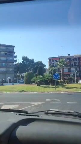 The video shows a view from inside a vehicle, looking out through the windshield. The scene is set in Cunit, Spain, on a bright, sunny day.

In the foreground, the dashboard of the car is visible, indicating the perspective of a driver or passenger.

The middle ground features a road with a roundabout. A person on a white dirt bike with black accents is riding on the road to the left of the roundabout. A blue circular traffic sign with three white arrows indicating a roundabout is prominently displayed on a pole. There are also grassy areas with palm trees and bushes surrounding the roundabout.

In the background, there are multi-story apartment buildings with balconies. The architecture suggests a Mediterranean setting. Further down the road, a white van and other vehicles are parked or driving. Streetlights and a pedestrian crossing signal are also visible.

Several signs are present. One sign has white text on a dark background and indicates directions to "Vilanova i la Geltrú" and "Barcelona" with road numbers "C-31" next to each. Other signs include yellow rectangular signs with text that appears to say "BON TOUR" and a blue square sign with a white pedestrian symbol and arrows. There are also flags on poles.

The overall impression is a snapshot of a sunny day in a residential area of a coastal Spanish town, with traffic and everyday street life. The clear blue sky and bright sunlight suggest it is daytime, likely mid-morning or afternoon.