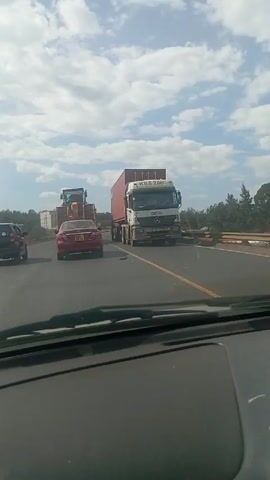 The video shows a view from inside a car moving on a highway behind a red sedan. A large truck with a red container and a blue construction vehicle are also visible in the distance ahead. Dust or debris is kicked up from the road as the car moves forward. The sky is visible above with scattered clouds. A malfunction in the video causes horizontal static lines and color distortion to flash across the screen throughout the duration of the clip.