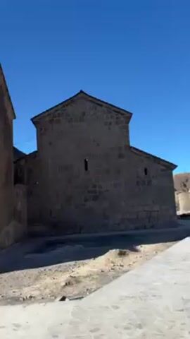 The video shows the exterior of a stone building complex under a bright blue sky. The buildings are constructed from dark gray stones, and their style appears to be medieval or historic. One building has a dome-shaped roof topped with a cross. The area around the buildings is partly paved with stone slabs, and there is some gravel or dirt visible. A child's voice can be heard.