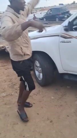The video shows a group of people and vehicles gathered near a makeshift wooden bridge in a sandy area. A white pickup truck is positioned near the entrance of the bridge. A man in a tan jacket and black shorts appears to be directing traffic. Other vehicles, including a dark SUV and a car with items strapped to the roof, are also present. People are seen on the bridge, some possibly in boats or canoes. The environment suggests a river or body of water, with a cloudy sky overhead.