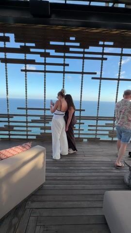 The video shows two women standing on a wooden deck looking out at the ocean. One woman is wearing a white swimsuit with white pants, and the other is wearing a brown dress. The deck has a decorative wooden structure. The ocean is visible through the structure and is a clear blue color. There are some white waves breaking on the shore. The sky is blue with some clouds. A man in a floral shirt and shorts is walking past the women on the right.