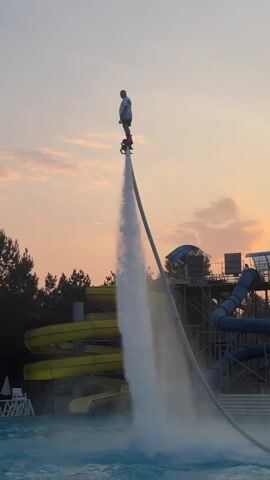 The video shows a person on a flyboard suspended in the air over a body of water. The flyboard is propelled by powerful jets of water, creating a dramatic spray that shoots upwards. The person, wearing a light-colored shirt and dark shorts, appears to be performing a maneuver, with their arms outstretched. Behind them, a water park with brightly colored slides is visible against a backdrop of trees and a sunset sky. The lighting suggests it is either sunrise or sunset, with warm hues coloring the clouds.