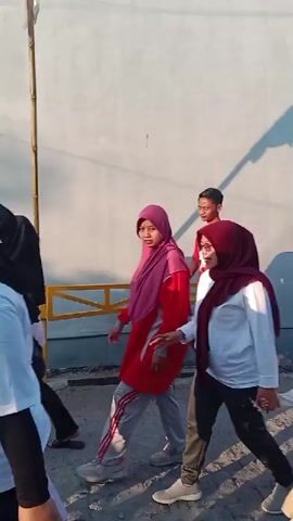 A group of people are walking together on a dirt path. The path is lined by a white wall on the left and buildings on the right. The sun is casting shadows on the wall. There are at least seven people visible in the foreground and background. In the foreground, on the left, a woman in a maroon hijab and glasses is holding hands with a child in a maroon hijab. To their right, a man in a white t-shirt and black baseball cap is walking and holding the child's other hand. Behind them, several other people are walking. Some are wearing hijabs and casual clothing. A red and white flag is visible on a pole on the right side of the frame. The lighting suggests it is daytime, possibly late afternoon due to the long shadows.