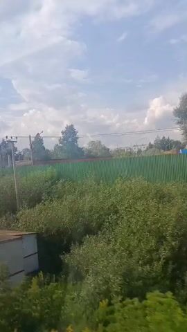 The video appears to be captured from a moving vehicle, likely a train, looking out a window. The view is dominated by lush green vegetation, including trees and bushes. In the foreground, there's a large, white, rectangular structure with a yellow triangular warning symbol on its side. To the left of this structure, a metal ladder or grate is visible. Overhead, there are power lines and utility poles. In the distance, a green corrugated fence runs horizontally across the frame, with more trees and some buildings visible behind it. The sky is partly cloudy with a bright blue sky peeking through. The overall impression is of an industrial or semi-rural area seen from a moving perspective.