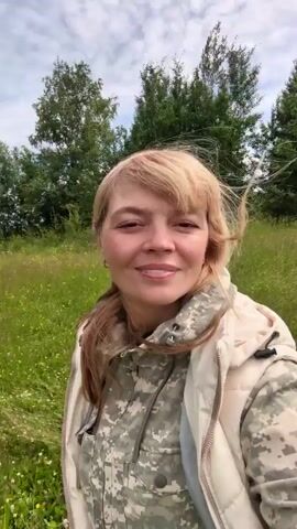 In this media file, a blonde woman is filmed in a close-up, selfie-style shot, walking through a grassy field with trees in the background. She is smiling and looking towards the camera. She is wearing a camouflage jacket and a light-colored vest. Her hair is blowing in the wind. The background shows a line of trees, possibly a forest, and a cloudy sky. The setting appears to be a rural or natural environment, likely during the daytime given the natural light. There is no visible text in the media. The overall mood appears to be cheerful and outdoorsy.