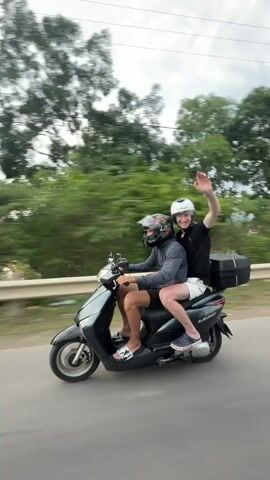 Two individuals, both wearing helmets, are riding on a dark-colored scooter. The person in front, appearing to be the driver, is wearing a grey hoodie, dark shorts, and sandals. The person behind, the passenger, is wearing a black t-shirt, light-colored shorts, and sneakers. They are both facing forward and appear to be interacting, with the passenger's arm around the driver. The scooter is moving on a paved road with a guardrail on the right side. Lush green trees and vegetation line the roadside, and the sky above is cloudy. The background suggests a rural or semi-rural environment. The video is captured from a side angle, following the scooter as it travels.