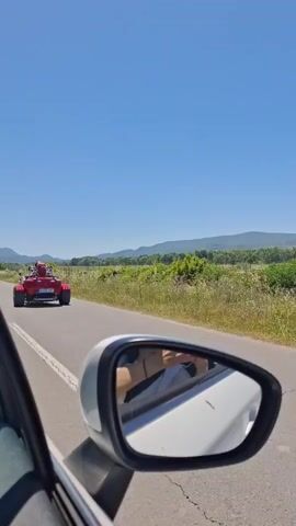 The video shows a car driving down a two-lane asphalt road on a sunny day. The focus is on a red three-wheeled vehicle in front, viewed through the driver's side mirror of the car.  The three-wheeled vehicle, similar to a trike, has two people riding in it, both wearing helmets. A large red backpack is strapped behind the passenger. The vehicle's license plate reads "AC MG 659". The surrounding environment includes green fields, shrubs, and distant hills under a clear blue sky. The road has a white painted line marking the lane.