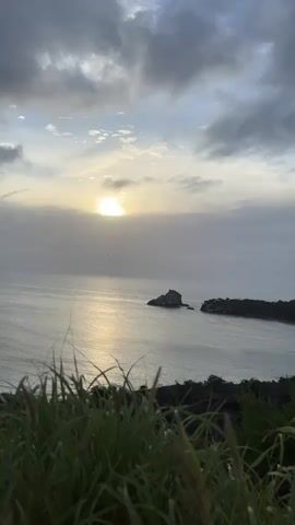 A video clip shows a serene ocean view at sunset. The sun peeks through a cloudy sky, casting a shimmering reflection on the water. Two small islands, one with a rocky outcrop and the other covered in vegetation, dot the sea. Lush green grass in the foreground frames the tranquil scene, its blades gently swaying as if in a soft breeze. The overall atmosphere is peaceful and calming, with a muted color palette dominated by grays, blues, and the warm glow of the setting sun. The video is likely filmed from a high vantage point, offering a panoramic vista of the coastal landscape.