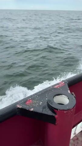 The video shows a view from a moving red boat on the open sea under a cloudy sky. White foam and wake are visible from the boat's movement through the dark grey water. In the distance, a large red gas carrier ship is visible on the horizon. Partially visible text on the side of the distant ship reads "GAS CARRI...". The scene depicts an overcast day at sea, likely offshore from Marina di Ravenna, Italy.