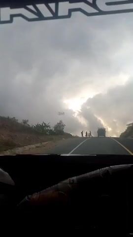 The video shows a view from inside a vehicle, looking out through the windshield at a road. The sky is cloudy and overcast, with the sun attempting to break through the clouds in the distance. On the side of the road, there are several people and bicycles. A truck is visible further down the road. The road itself is paved and has yellow lines marking the lanes. The foreground of the video shows the interior of the vehicle, including a steering wheel. The scene appears to be a rural or suburban area.