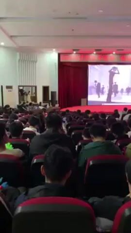 The scene shows a large auditorium filled with people seated in rows of red chairs. A person is on stage playing an electric guitar in front of a large screen displaying an animated figure in a black suit. The screen also displays Chinese characters at the top. The room is well-lit, and the audience members appear to be watching the performance. Some audience members are holding up their phones. There are decorative wall panels and doorways on the side of the auditorium.