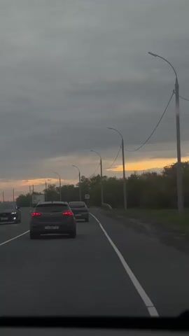 The video shows a view from inside a car driving on a highway. The camera is positioned behind a dark-colored car. The sky is overcast with a hint of orange from the setting sun visible on the horizon. There are street lights and power lines along the side of the road, as well as trees and greenery. There is at least one other vehicle visible in the distance.
