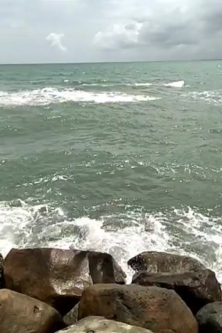 The video shows an ocean on a cloudy day, with large rocks in the foreground. The waves are crashing against the rocks, creating white foam. The water is a greenish-blue color, and the sky is overcast.