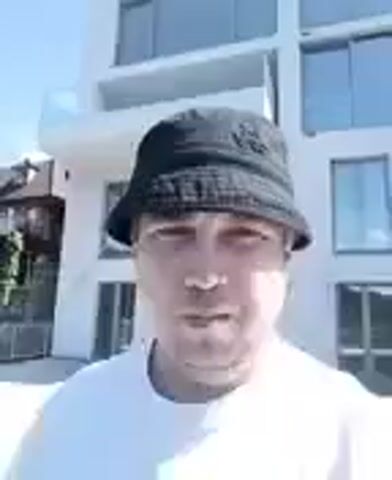 A person wearing a white shirt and a black bucket hat faces the camera, speaking. The person is outdoors, with trees and a building visible in the background. The sky is blue and there are some clouds.