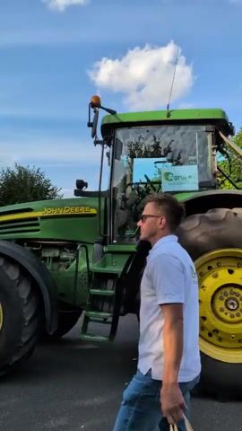 A man in a white polo shirt and black shorts walks past a large green John Deere tractor. The tractor has a trailer attached, which is decorated with green branches and colorful streamers. Several people, including children and adults, are riding on the trailer, smiling and waving. The trailer has a banner that reads "Agrar eg Guthmannshausen Moderne Landwirtschaft fur unsere Region." The scene appears to be an outdoor event, possibly a parade, under a bright blue sky with fluffy white clouds.