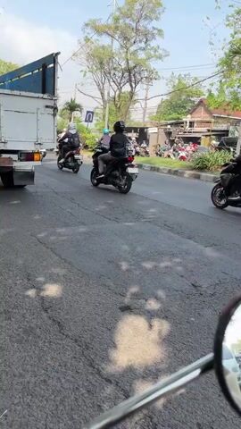 The video shows a street scene with several motorcycles. In the foreground, a motorcycle's rearview mirror is visible, reflecting a street with some cars. Ahead, several motorcycles are riding on the road, all carrying riders wearing helmets and black jackets. The motorcycles are in motion. On the right side of the road, there are trees and some buildings, possibly houses. The sky is clear and blue, suggesting daylight. The road appears to be paved and has lane markings. The overall impression is of a busy street with motorcycle traffic.
