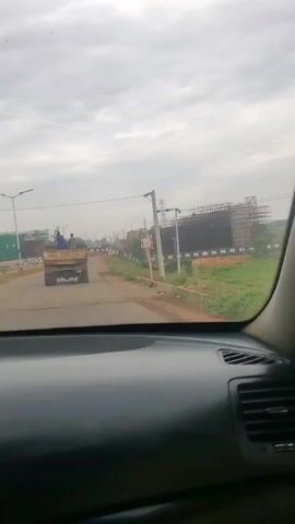 The video is shot from inside a car, looking out the windshield. The car is driving on a paved road in a rural area. Ahead, a truck with at least two people riding in the back is driving in the same direction. To the right of the road, a large construction site is visible. The sky is overcast. The camera briefly pans to the right, revealing a green field and a concrete wall. The video then returns to the view through the windshield.