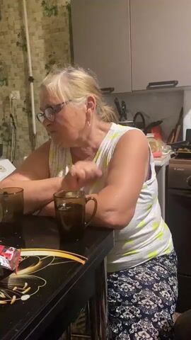 An older woman with blonde hair and glasses is sitting at a table in what appears to be a kitchen. She is wearing a white sleeveless top with a colorful pattern and a dark skirt with a floral print. She has her arms resting on the table, and there are two mugs in front of her. The woman looks towards the camera and smiles. There are some items on the table, including what looks like a package of something red and white. In the background, kitchen cabinets are visible.