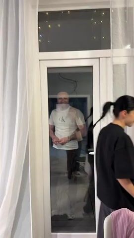 The video shows a woman taking a selfie in a large mirror, reflecting a room undergoing renovation. The woman has long dark hair, wears a light beige jacket, a grey top, and jeans. A man in a white t-shirt and jeans is partially visible in the left side of the reflection.  A small child is visible in the bottom right corner of the mirror. The man leans into the frame, looking at the camera. The room behind the mirror appears to be under construction, with visible wires and unfinished walls. The video has a casual, home-shot feel.