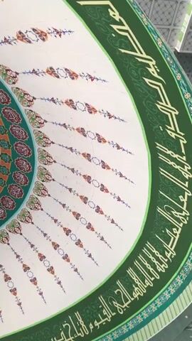 The camera is pointing upwards at the ornately decorated ceiling of a structure. A circular design dominates the center, featuring concentric rings of blue, gold, and green, filled with intricate patterns and Arabic script. This central design is surrounded by a ring of green floral patterns, interspersed with designs containing more script. Radiating outward from this central pattern are teardrop-shaped designs that contain Arabic script and floral decorations. The entire design is set against a white background and bordered by bands of green script. A chandelier hangs from the center of the ceiling.