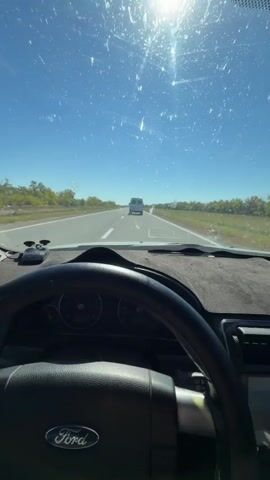 The video shows a view from inside a car looking out the windshield on a sunny day. The car is on a paved road with at least two lanes in each direction. The road is surrounded by sparse vegetation and trees. In the distance, a white van is visible driving in the same direction as the camera car. The windshield has a significant amount of dust and smudges, and there are visible scratches. The sun is bright and appears high in the sky. The interior of the car is visible in the foreground, including the steering wheel with a Ford logo, the dashboard, and a rearview mirror with a beaded string hanging from it. A black dash mat covers the dashboard. The overall impression is of a drive through a rural or semi-rural area.