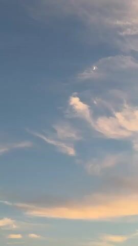 A video of a crescent moon in a pale blue sky with wispy clouds.