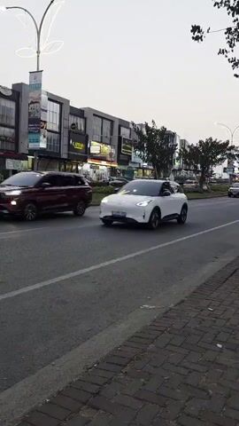 The video shows a white electric car driving on a street. The car is moving from left to right in the frame. The car has a modern design with sleek lines. The background shows buildings and trees. The sky is overcast. The video is shot from a slightly elevated position, possibly from a sidewalk or a building. There is another dark-colored car on the left side of the frame, also moving in the same direction. The street appears to be a multi-lane road. There is also a motorcycle visible in the distance, also moving in the same direction. The overall atmosphere seems to be that of a busy urban environment.