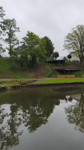 The video shows a tranquil scene by a body of water, likely a lake or canal. The water's surface reflects the overcast sky and the lush green trees lining the bank. On the bank, there are two elevated wooden structures, possibly viewing platforms or covered seating areas, with stairs leading up to them. Below these, a paved path runs along the water's edge, bordered by a strip of bright green grass and wooden benches. Further along the bank, several trees stand tall, their leaves appearing full and healthy. In the distance, a fountain sprays water high into the air, creating a refreshing visual contrast to the muted tones of the cloudy sky. Some aquatic plants, possibly lily pads, are visible in the foreground of the water. The overall impression is one of a peaceful, well-maintained park or recreational area.