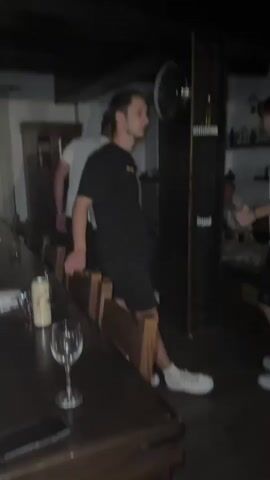 The video shows a dimly lit indoor space, possibly a bar or a party venue. Several young adults are present. In the foreground, a person in a black t-shirt and shorts is leaning on a wooden bench. To their right, another person in a white t-shirt and black shorts is standing. Further back, a person in a patterned blue and white shirt and white pants is visible. In the background, there are shelves with bottles, a clock on the wall, and a couch with people seated on it. There is a speaker with colorful lights on the right side of the frame. The overall atmosphere appears to be social and relaxed.