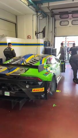 The video shows a busy pit garage. The focus is on a green and blue race car, likely a Lamborghini, with the number 63 visible on its rear. Several people, dressed in racing team attire, are working on the car. Some are standing, observing, while others are bent over, appearing to be making adjustments or repairs. One person is near the rear of the car with tools. Another person is to the right, also working on a car. In the background, more team members are visible, and the garage door is open, revealing daylight and possibly other cars or structures outside. The environment is utilitarian, with exposed ceilings, lighting fixtures, and a red, durable floor. There is a general sense of activity and focused work within the racing team's preparation area.