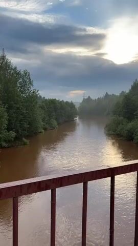A drone captures a view of a wide, muddy brown river flowing through a forest. The water is calm with gentle ripples. On the right bank, a small, long, wooden boat is tied to a tree, partially submerged in the murky water. The sky is overcast with some sunlight breaking through the clouds. The scene is viewed from above, with a red railing in the foreground, suggesting the shot is taken from a bridge or elevated platform.