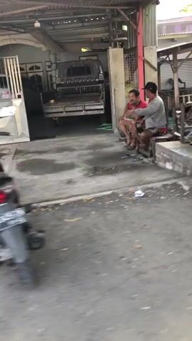 The video opens with a close-up of a motorcycle in motion on a paved street. The camera then pans to the right, revealing two men sitting on a concrete ledge in front of a building. The building appears to be a workshop or garage, with a pickup truck parked inside. The man on the left is wearing a red t-shirt and the man on the right is wearing a gray long-sleeved shirt and a red baseball cap. Both men are wearing shorts. They appear to be in conversation, with the man on the right gesturing with his hands. The environment is somewhat cluttered, with various tools and materials visible in the background. The lighting suggests it is daytime.