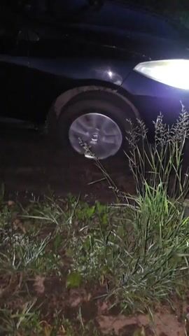 A black car is stuck in mud. The camera pans across the car's front wheel and fender, showing how deeply it is embedded in the thick, dark mud. The surrounding area is overgrown with grass and other vegetation, and the lighting suggests it is either dusk or nighttime. The car's headlights are on, casting a bright beam into the scene. There are no people visible in the video. The overall impression is of a vehicle that has become mired in difficult terrain.