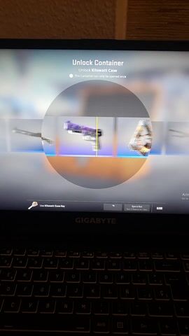 The video displays a laptop screen showing a video game interface, likely from Counter-Strike. The laptop itself is black with a "GIGABYTE" logo visible below the screen on the right side. A black keyboard with white lettering and a Spanish layout (indicated by keys like "Ñ") is visible on the right.

The screen shows an "Unlock Container" interface. In the center, there's a circular spinner displaying various weapon skins, representing items from a "Kilowatt case." The text "Unlock Kilowatt case" and "This container can only be opened once" are visible on the left side of the screen. Below these, there's a button with a key icon and the text "Use Kilowatt Case Key."

As the video progresses, the spinner rotates rapidly, showing different weapon skins. A yellow line acts as a pointer in the center of the circle. The spinner then slows down and eventually stops.

The final item revealed is a shotgun skin. The screen updates to show a full-screen view of the shotgun, identified as "Nova | Dark Sigil" from "The Kilowatt Collection." A description of the weapon skin appears below its name. The text reads: "The Nova's rock bottom price tag makes it a great ambush weapon for break-strapped teams. It has been custom painted with longships and dragons. It emerged from the fog of war." A "CLOSE" button is visible in the top right corner of the screen.