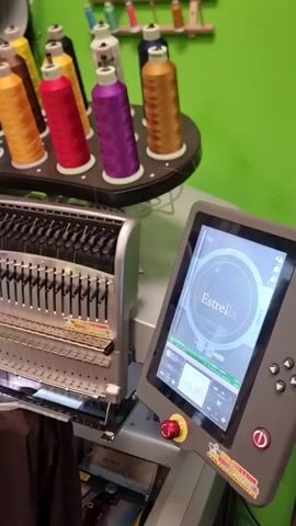 The media file displays an industrial embroidery machine in operation, likely in a workshop or studio. The primary subject is the embroidery machine itself, a complex piece of equipment with multiple needles and spools of colorful thread. A digital touchscreen interface is visible, displaying the word "Estrella" within a circular design, indicating the embroidery pattern being executed. The spools of thread are arranged on a tiered rack, showcasing a vibrant selection of colors including yellow, red, purple, and gold.

The machine is actively embroidering the name "Estrella" onto a dark fabric, which is held taut within a hoop. The needles are in motion, creating the stitched design. The background is a bright, almost fluorescent green, suggesting a painted wall or backdrop, which contrasts sharply with the metallic sheen of the machine and the rich colors of the threads. The overall impression is one of a professional setting where custom embroidery is produced.

While the location is contextually given as Tullamore, Ireland, there are no specific visual cues within the media file itself to confirm this. The lighting appears to be artificial, consistent with an indoor workspace, and there are no indications of the time of day or weather conditions. The visible text, "Estrella," is part of the embroidery design on the screen and the fabric. Another piece of visible text, "RUSTYPATCH.COM," is present on a sticker on the control panel, likely indicating the manufacturer or service provider of the embroidery machine. No people are visible in the media file.