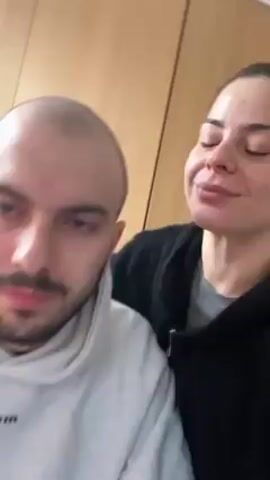 A woman is kissing a man on the cheek in this video. The man has a shaved head and a short beard, and he is wearing a grey hoodie. The woman has her hair pulled back and is wearing a black hoodie. They are standing indoors in front of a light-colored wooden wall. The woman is making kissing sounds as she kisses the man. The camera is close to their faces.