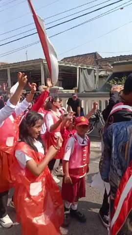 A group of people, mostly children and adults, are participating in an outdoor parade. They are dressed in red and white attire, with many wearing red plastic aprons over their clothing. Some individuals are holding a long red and white flag or banner, which is partially visible at the top of the frame. The people appear to be in motion, with some raising their hands in the air and seemingly dancing or cheering. The environment suggests a street or road in a residential area, with buildings visible in the background and overhead power lines. The lighting indicates it is daytime, likely sunny. One child in the foreground is holding a small device, possibly a camera or phone, and aiming it towards the front. The overall atmosphere seems celebratory and festive.