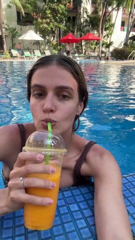 A woman in a brown swimsuit is in a swimming pool. She is holding a clear plastic cup filled with an orange liquid, presumably a juice or smoothie, and drinks from it with a green straw. The pool's edge is visible in the foreground with blue tiles. In the background, there are lounge chairs with white umbrellas and some red umbrellas, along with palm trees and buildings. The woman looks at the camera, pauses drinking to make a face and looks forward.