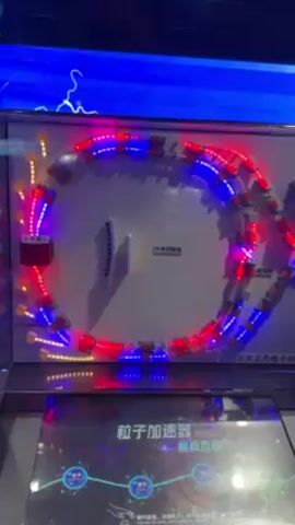 The video shows a model of a particle accelerator. The model features a circular track illuminated with alternating red and blue lights. Small, square elements are placed along the track, presumably representing magnets or accelerating cavities. A section on the left-hand side of the track is lit with yellow lights, perhaps representing the particle source. The model has labels written in Chinese characters. The video is recorded indoors, under artificial lighting. The background includes a blue wall or screen with a depiction of lightning. The video appears to be shot from a slightly low angle. The base of the model is also visible, featuring a screen displaying diagrams and text, also in Chinese. The text on the base identifies the model as a particle accelerator. The scene appears to be in a museum or science center.