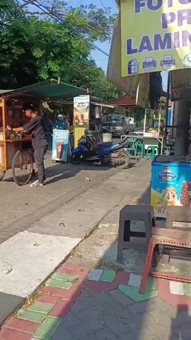 The video shows a street scene with several businesses. On the right, there is a stall with a large yellow sign that reads "FOTOCOPY PRINT LAMINATING" in large purple letters, along with a phone number. Next to this stall, there is a blue cabinet or kiosk with a sign that says "FOTOCOPY PRINT CETAK FOTO." In front of the blue kiosk is a blue ice cream freezer displaying various ice cream products. To the left of the photocopy stall, there are several other small stalls or shops with awnings. One stall has a sign with "2500" on it, possibly indicating a price. A person wearing a black shirt and pants is seen walking away from a food cart on the left side of the frame. A blue motorcycle is parked in front of one of the stalls. The ground is paved with concrete and decorative tiles. Trees are visible in the background, suggesting an outdoor environment. The sun is shining, casting shadows.