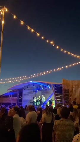 The video shows a nighttime outdoor concert or festival. In the background, a band is performing on a stage illuminated by colorful lights, which shift between blue, red, and green. String lights hang across the scene, adding to the festive atmosphere. In the foreground, a crowd of people are gathered, facing the stage and watching the performance. The sky is dark blue, indicating it is evening or night. The overall impression is of a lively outdoor event.