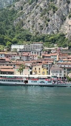 A wide shot captures a picturesque Italian village nestled at the base of a steep, rocky mountain overgrown with trees. The village features a dense cluster of buildings with terracotta roofs, some painted in warm, muted colors. In the foreground, a large, white paddlewheel boat with multiple decks cruises across a calm, turquoise body of water. People are visible on the decks of the boat and along the waterfront of the village, where outdoor seating areas with green awnings are set up. The scene is bathed in bright sunlight, indicating a clear day. The boat is moving from left to right across the frame.