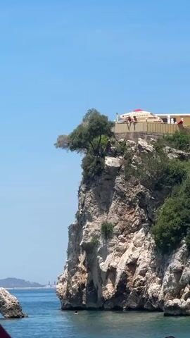 The video captures a scenic coastal cliffside on a bright, sunny day with a clear blue sky. The cliff is rugged and imposing, covered in patches of greenery and rocks. Atop the cliff, a structure resembling a restaurant or cafe with a yellow facade and a red roof is visible, with several people standing near the edge.

The main action centers around a person who is captured mid-air, leaping from a considerable height off the cliff face into the turquoise water below. A splash erupts where they hit the water, creating a dynamic visual moment. Further out in the water, another person can be seen swimming or floating. In the background, the coastline stretches out, with hints of distant buildings and islands, suggesting a popular tourist destination. The overall atmosphere is one of adventure and exhilaration, likely a summer day activity.