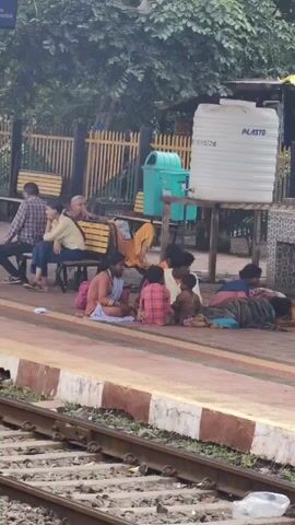 A group of people are sitting and lying on a train platform. In the foreground, a group of people, including women and children, are sitting on the ground near the train tracks. Some are interacting with each other. In the background, other people are sitting on benches and one person is lying down. There is a large white water tank and a green trash bin visible. The scene appears to be at a train station.